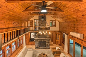 A spacious log cabin interior with a stone fireplace, wooden beams, and a balcony overlooking a deck. - Hilltop Lackawaxen Gem w/ River View & Fire Pit (Lackawaxen)