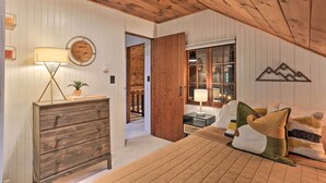 Un dormitorio con techo de madera, un aparador, una cama con almohadas, una mesita de noche con lámpara y una ventana con vistas al exterior.