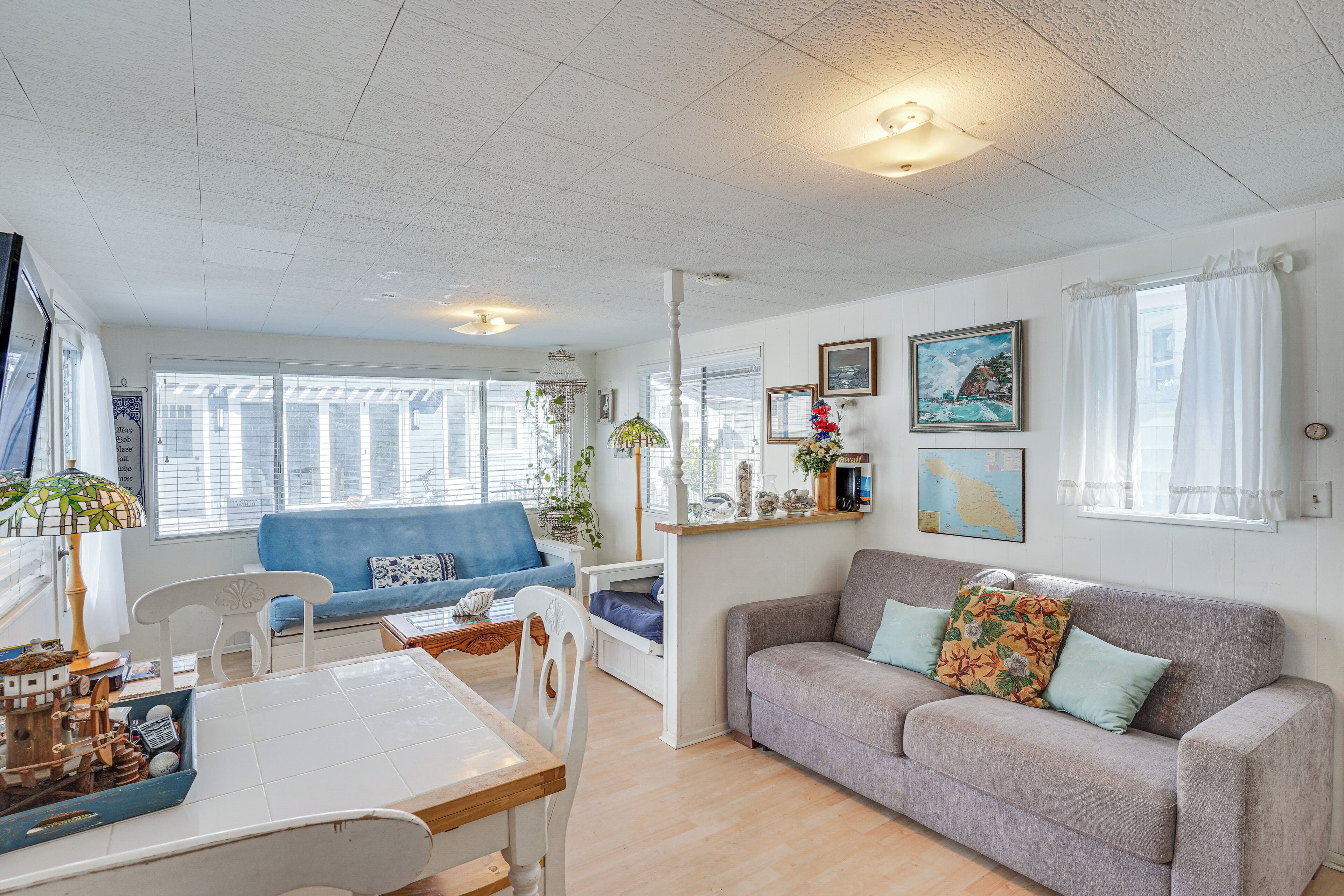 Ein großzügiger Wohnbereich mit blauem Sofa, grauem Sofa mit floralen Kissen, Esstisch und Stühlen sowie weißer Decke mit Rasterleuchten.