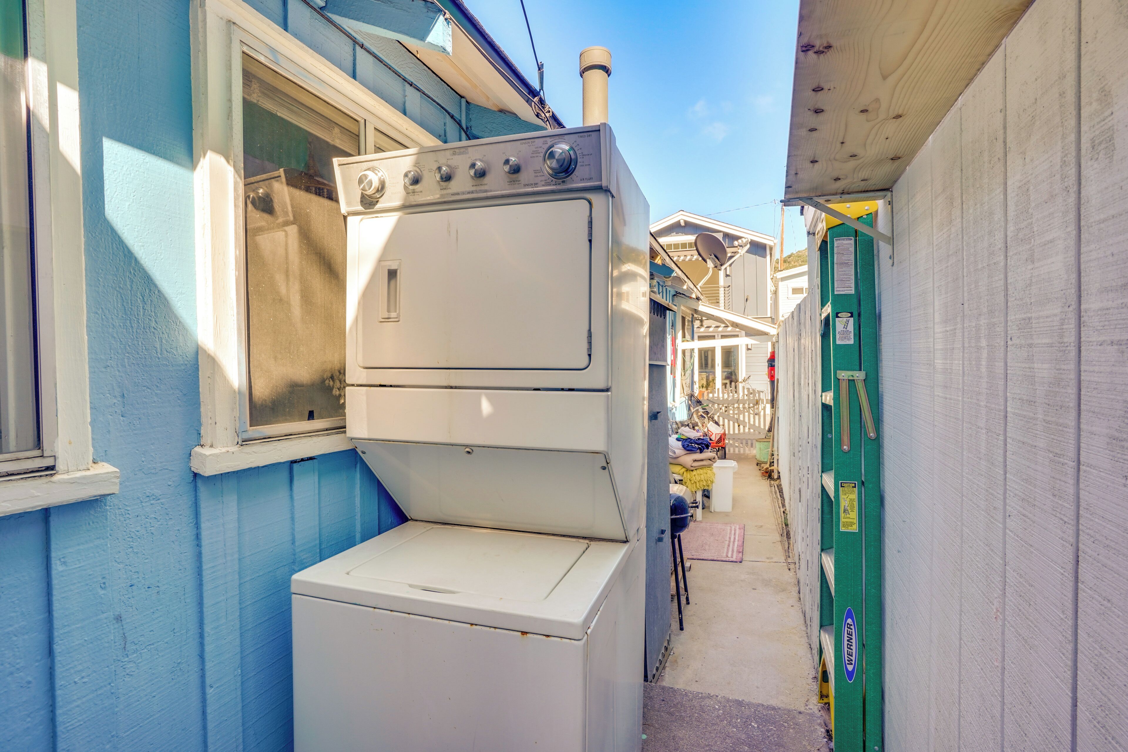 Une ruelle étroite où l’on aperçoit une machine à laver et un sèche-linge blancs, placés l’un sur l’autre, adossés à un bâtiment bleu.