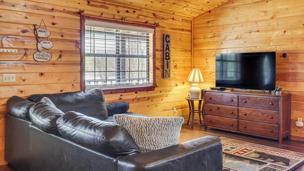 A cozy cabin interior with a leather sofa, wooden walls, a television, and a window with blinds. - Broken Bow 'starlight Cabin' w/ Private Hot Tub! (Broken Bow)