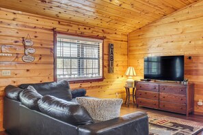 A cozy cabin interior with a leather sofa, a wooden cabinet, a television, and a window with blinds. - Broken Bow 'starlight Cabin' w/ Private Hot Tub! (Broken Bow)