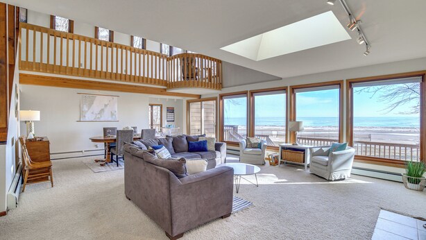 Ein geräumiges Wohnzimmer mit hoher Decke, großen Fenstern und einem Balkon mit Blick auf den Strand.