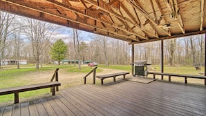 A covered wooden deck with benches and a grill area, overlooking a grassy area with trees and a distant building. - Rustic & Modern Russell Cabin w/ Grill + Deck (Russell)