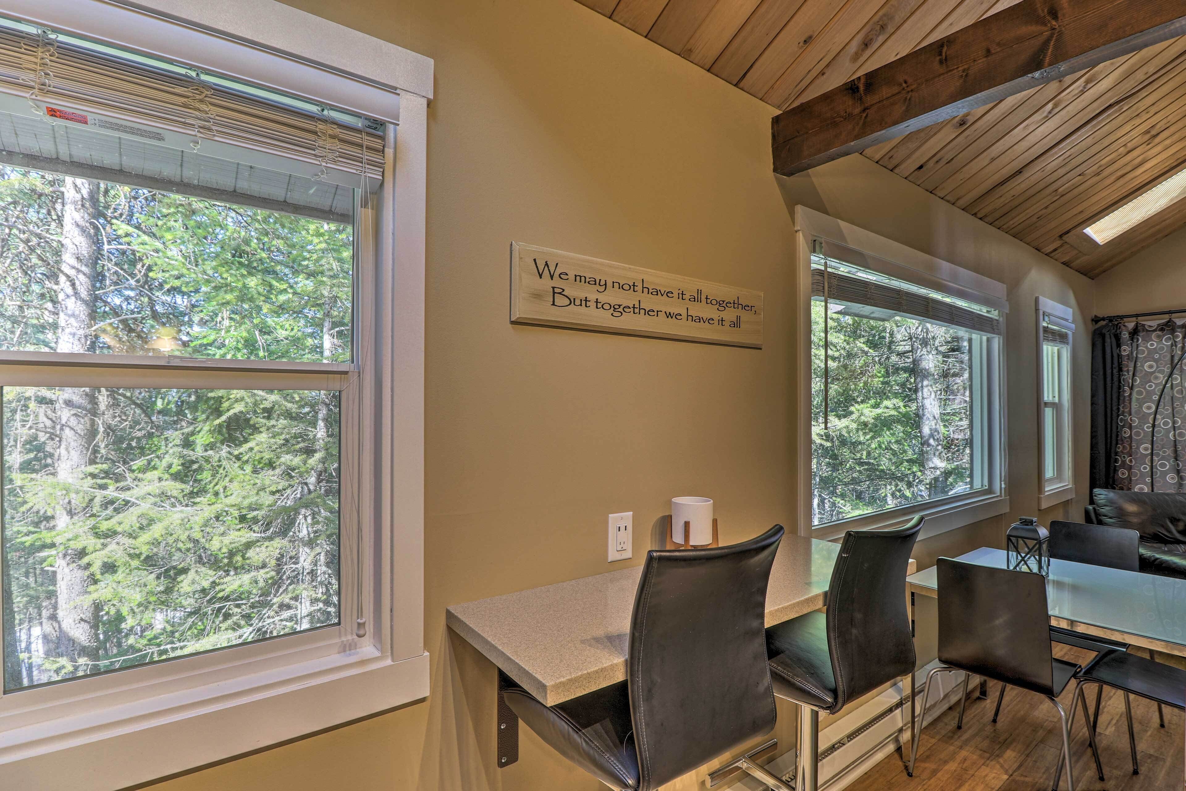A dining area with a table and chairs, large windows with a view of trees, and a motivational sign on the wall.