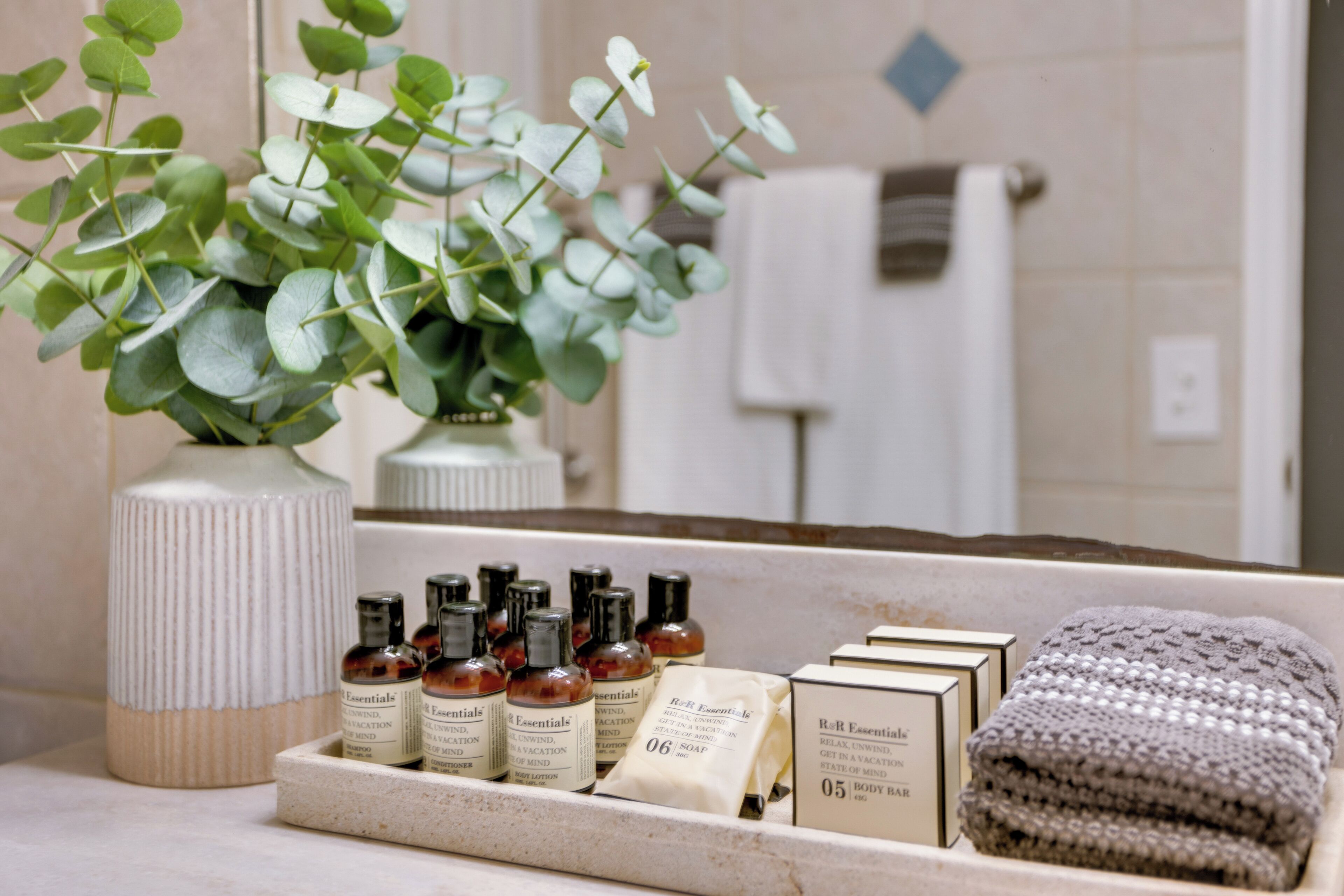 A bathroom vanity with toiletries, a vase of green leaves, and a towel.