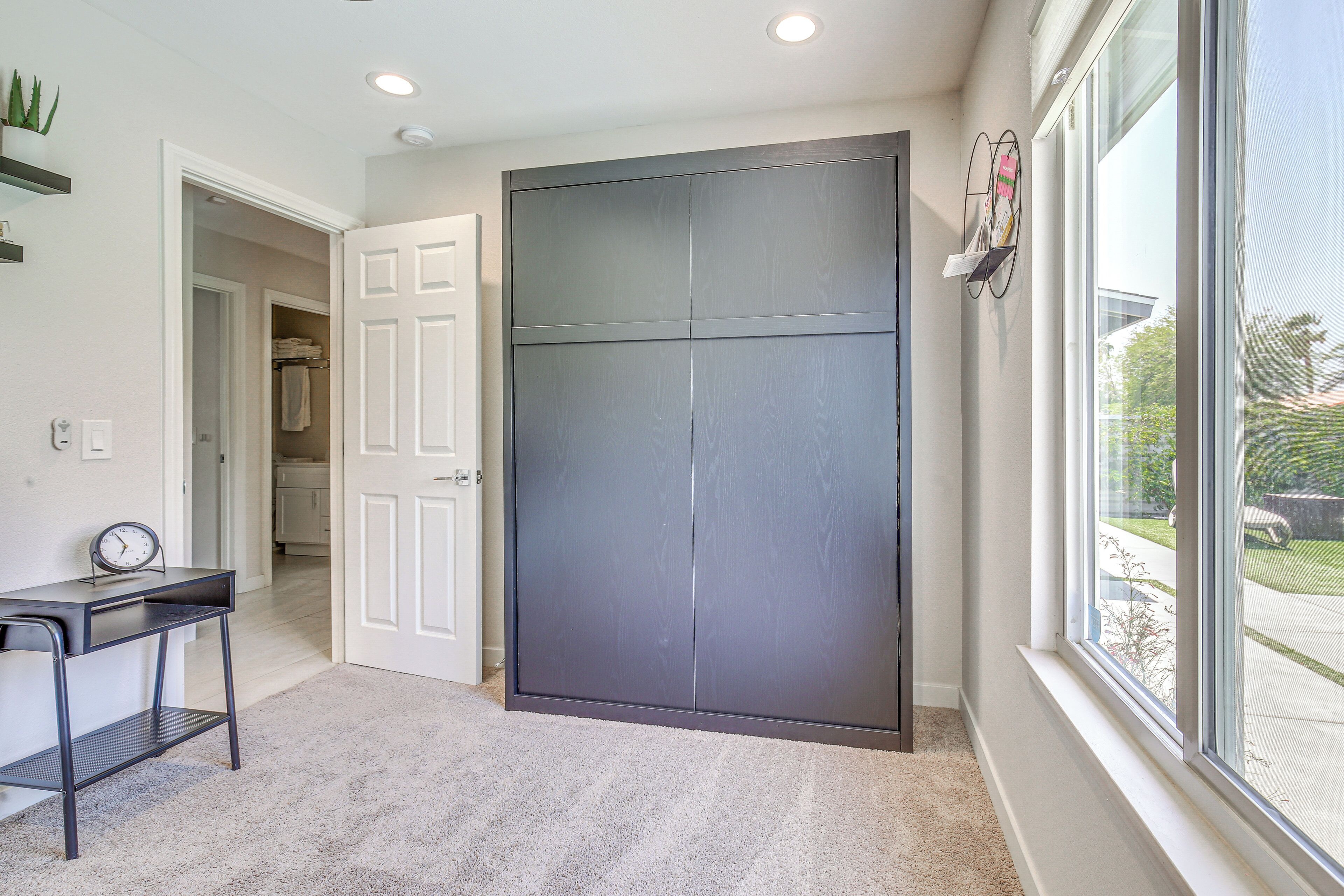 A hallway with a large sliding door, a small table with a clock, and a window with a view of the outside.