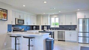 A modern kitchen with white cabinets, stainless steel appliances, a central island with bar stools, and a large window providing natural light. - Palm Springs Pad w/ Outdoor Kitchen + Views! (Palm Springs)