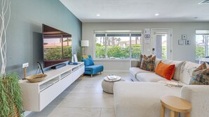A modern living room with a flat-screen TV, a sofa, a blue armchair, and a large window offering a view of greenery. - Palm Springs Pad w/ Outdoor Kitchen + Views! (Palm Springs)