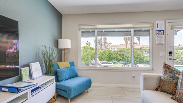 Ein Zimmer mit Schreibtisch, zwei Sofas, einem Fernseher und einem Fenster mit Blick auf Palmen und einen Pool.
