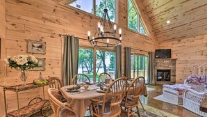 L’intérieur d’un chalet en bois présente une table à manger dressée pour quatre personnes, entourée de chaises en osier, d’un lustre et d’une cheminée.