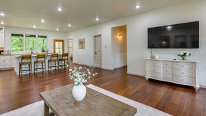 A spacious living area with a wooden coffee table, a dining table with chairs, a white cabinet, and a flat-screen TV mounted on the wall.