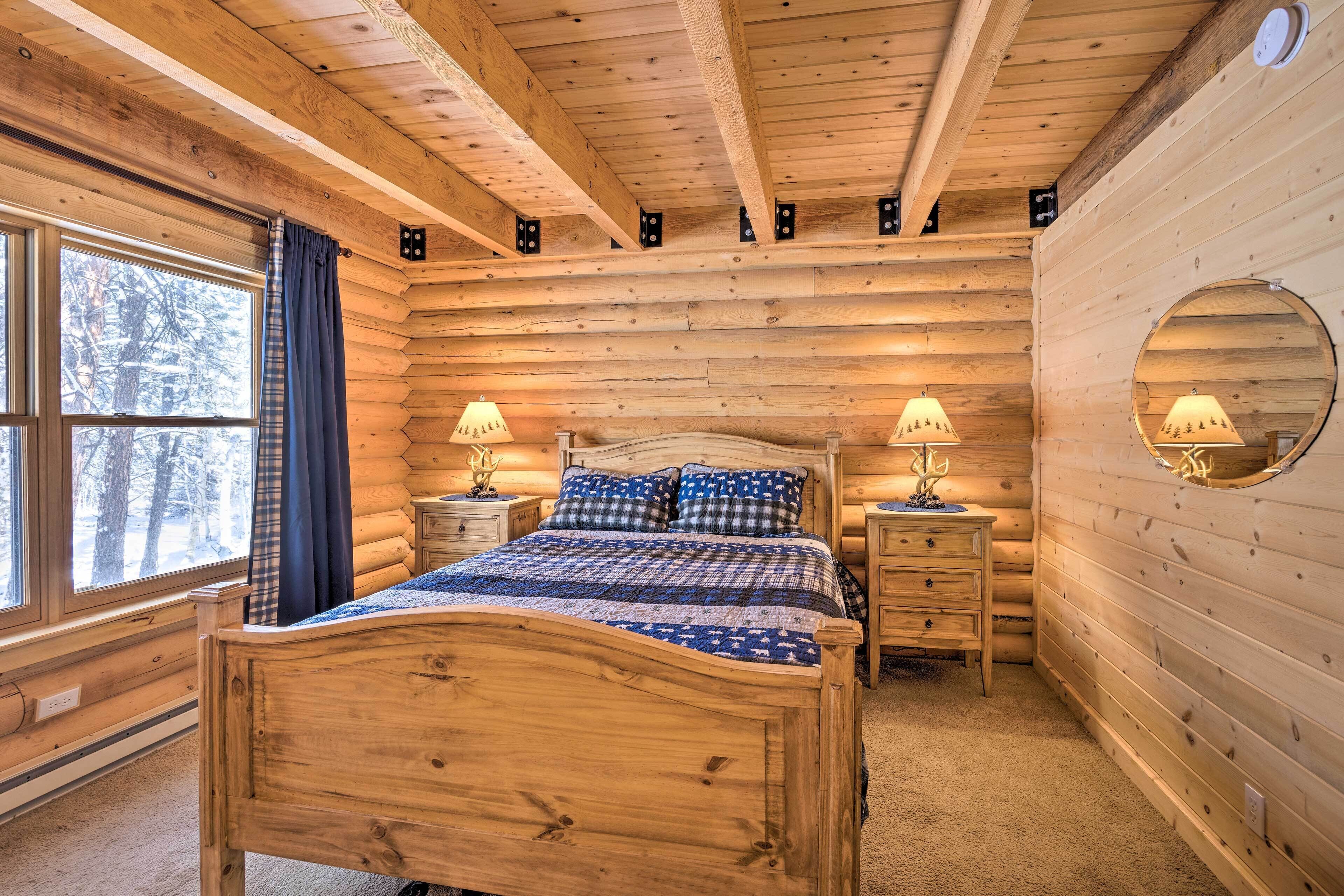 A log cabin bedroom with a wooden bed, two bedside tables with lamps, a window with curtains, and a circular mirror on the wall.