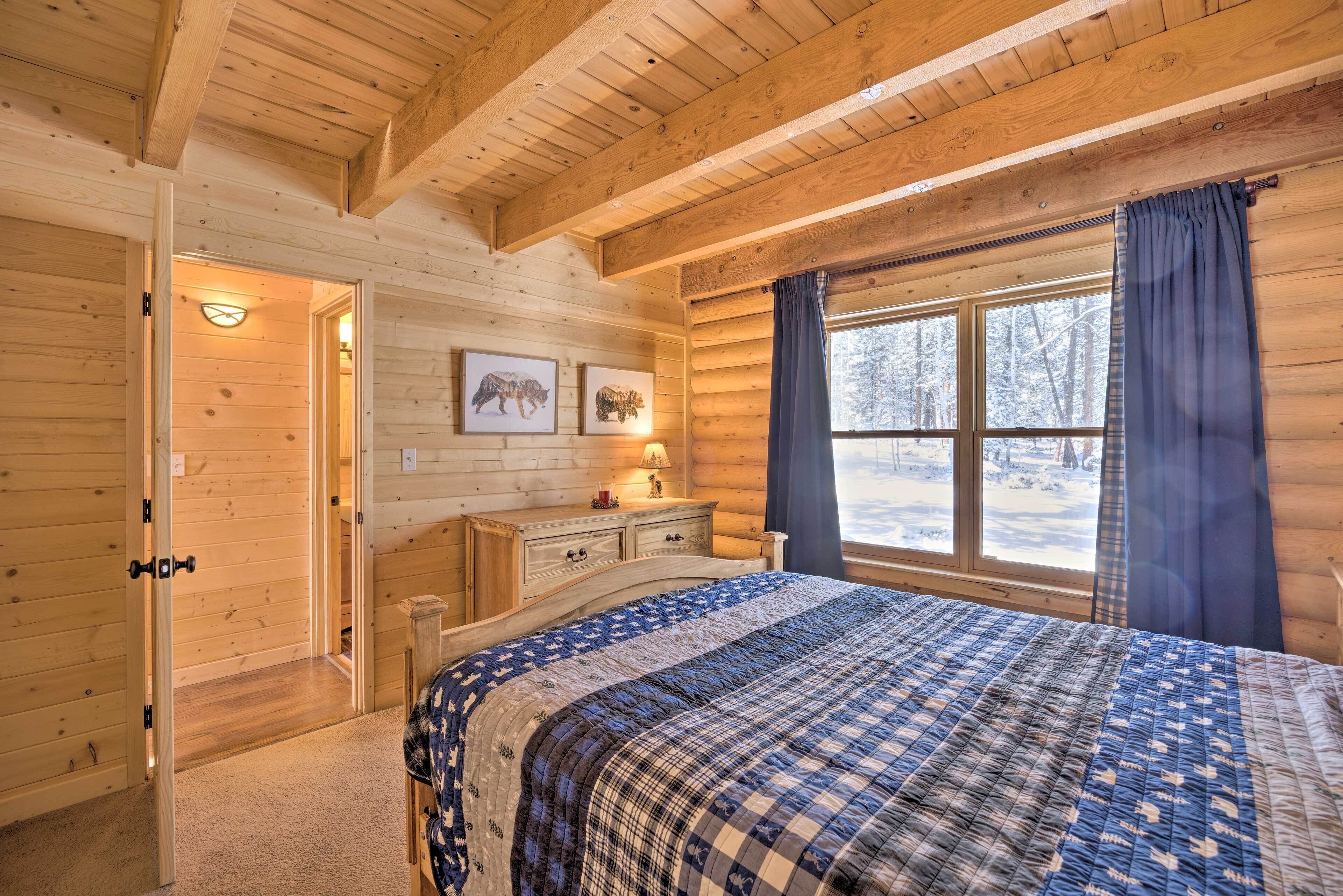 Ein Schlafzimmer in einer Blockhütte mit Bett, Holzwänden, einem Schminktisch, einem Fenster mit Vorhängen und einer Wandleuchte.