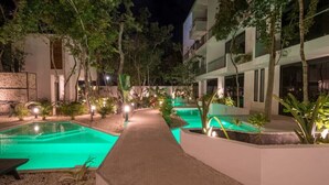 Una vista notturna di un cortile alberghiero con due piscine, circondato da alberi e illuminato da luci lungo il percorso.