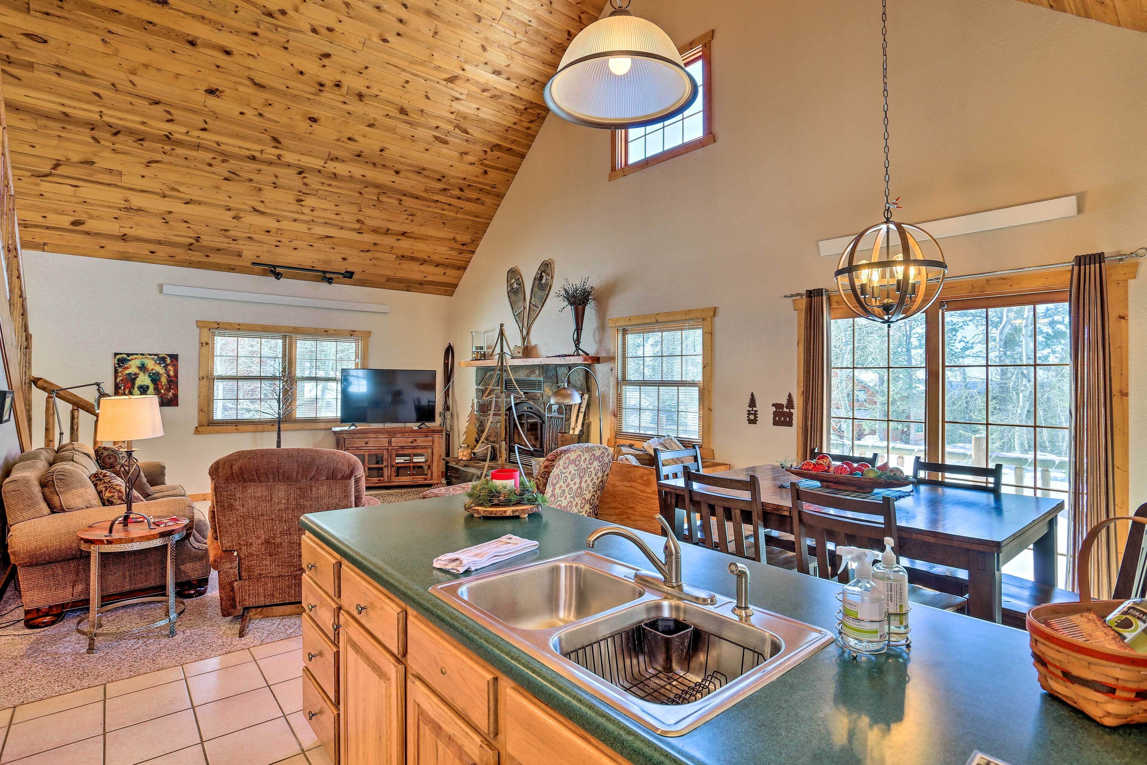 A cozy cabin interior with a wooden ceiling, a dining area, a kitchen with a large island, and a living room with a sofa and a lamp.