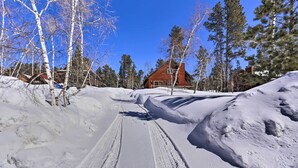 En snötäckt stig leder fram till en stuga av trä med ett rött tak, omgiven av träd och snövallar.
