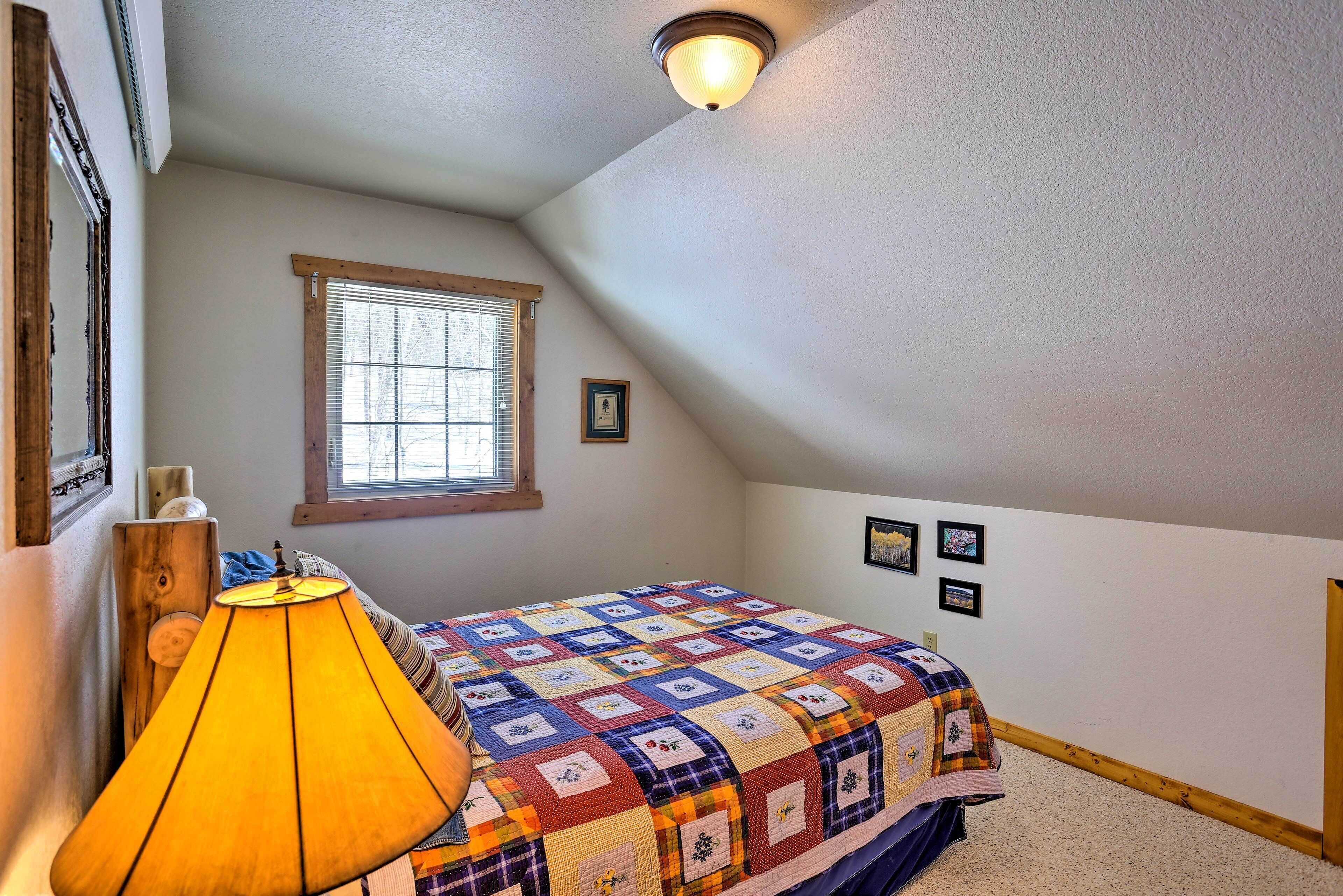 A bedroom with a sloped ceiling, a window with blinds, a colorful quilt on the bed, a lamp, and a small picture on the wall.