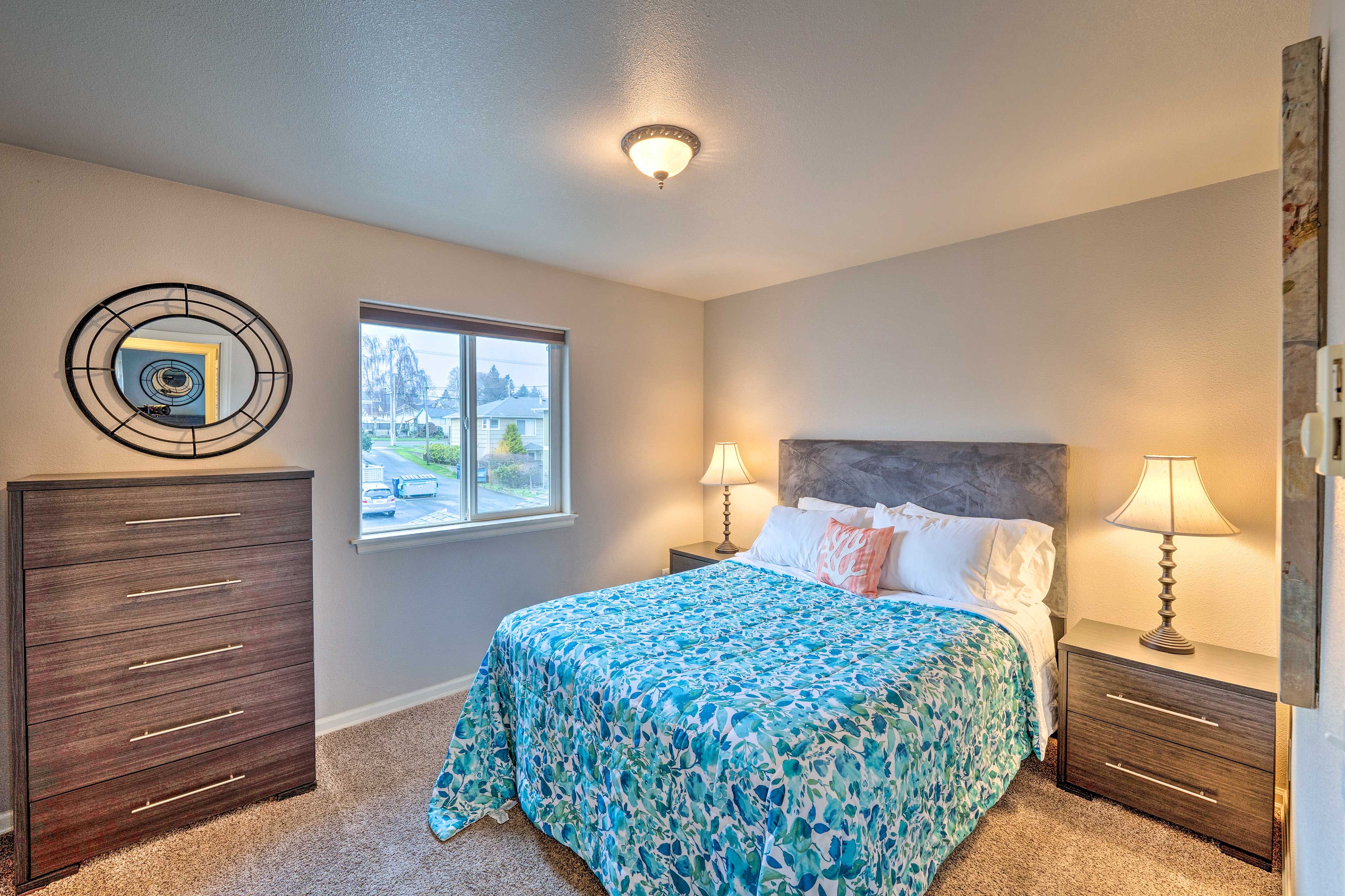 A bedroom with a bed, two bedside tables with lamps, a dresser, a window, and a wall-mounted mirror.