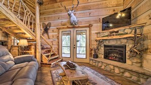 A log cabin interior with a stone fireplace, a mounted deer head, a sofa, and a staircase. - Stunning Creekside Cosby Cabin w/ Deck + Fire Pit! (Cosby)