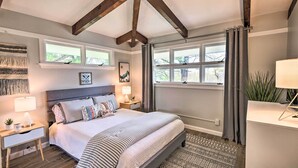 Ein Schlafzimmer mit einem großen Bett, zwei Nachttischen, einem kleinen Tischchen, einem Fenster mit Vorhängen und einer Decke mit freiliegenden Balken.