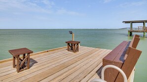 A wooden deck with a bench and a small table, overlooking calm water and a distant bridge. - Waterfront Port Isabel Family Home w/ Pool & Pier! (Port Isabel)