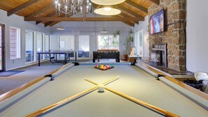 A pool table with four cues and a white ball in the center, a foosball table, a fireplace, and a television mounted on a stone wall. - Lincoln City Vacation Rental w/ Pool & Ocean Views (Gleneden Beach)