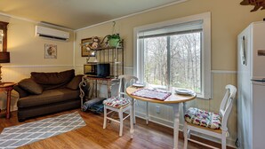 Un salon chaleureux avec un canapé marron, une table basse en bois et une chaise blanche. Il y a une fenêtre donnant sur des arbres, un climatiseur et une petite étagère avec des objets décoratifs.