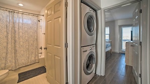 A laundry room with a front-loading washer and dryer, a shower with a patterned curtain, and a hallway leading to a bedroom. - Mexico Beach Home w/ Balcony - Steps to Beach (Port St. Joe)