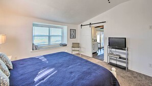 Quarto com uma cama grande, televisão sobre suporte, ventilador de teto, janela com persianas e porta para outro cômodo.