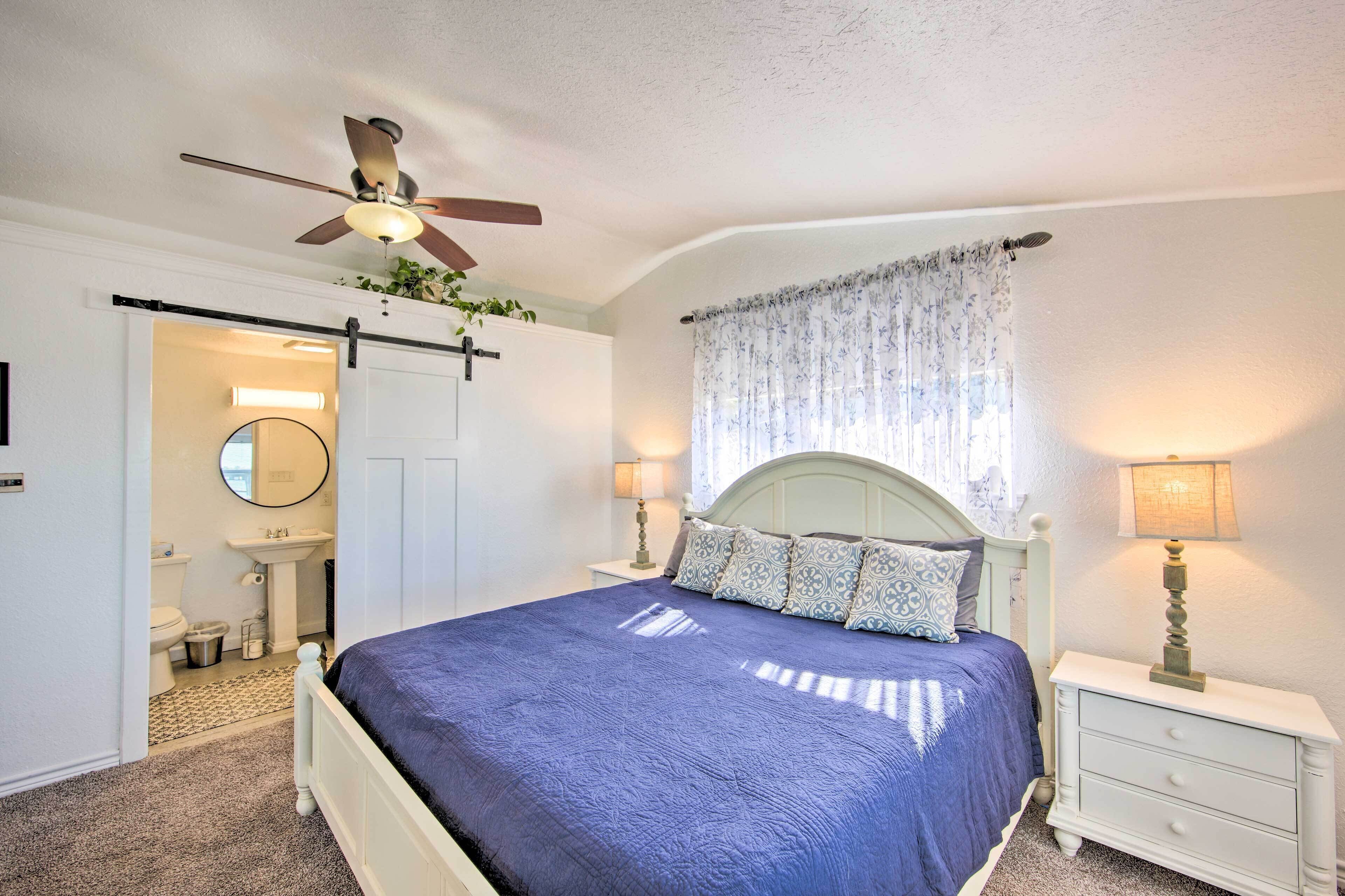 Une chambre à coucher avec un lit, un ventilateur de plafond, une table de chevet, une lampe, une fenêtre avec des rideaux et une salle de bain visible à travers une porte ouverte.