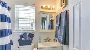 A bathroom with a toilet, sink, and mirror. There is a window with blinds, a towel rack, and a basket of towels. - Steps to Surfside Beach With Easy Ramp Access! (Freeport)