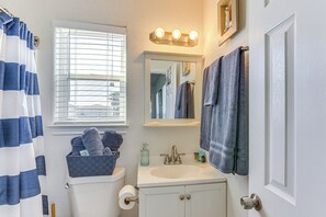 A bathroom with a toilet, sink, and mirror. There is a window with blinds, a towel rack, and a basket of towels. - Steps to Surfside Beach With Easy Ramp Access! (Freeport)