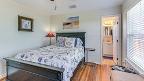 A bedroom with a bed, a ceiling fan, a nightstand, a window with blinds, a door leading to a bathroom, and a framed picture on the wall. - Steps to Surfside Beach With Easy Ramp Access! (Freeport)