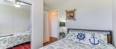 A bedroom with a bed featuring nautical-themed bedding, a ceiling fan, and a small bedside table with a lamp.