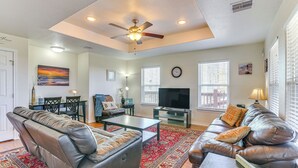 A spacious living room with a large sofa, a coffee table, and a television. There is a ceiling fan, a rug, and a painting on the wall. - Steps to Surfside Beach With Easy Ramp Access! (Freeport)