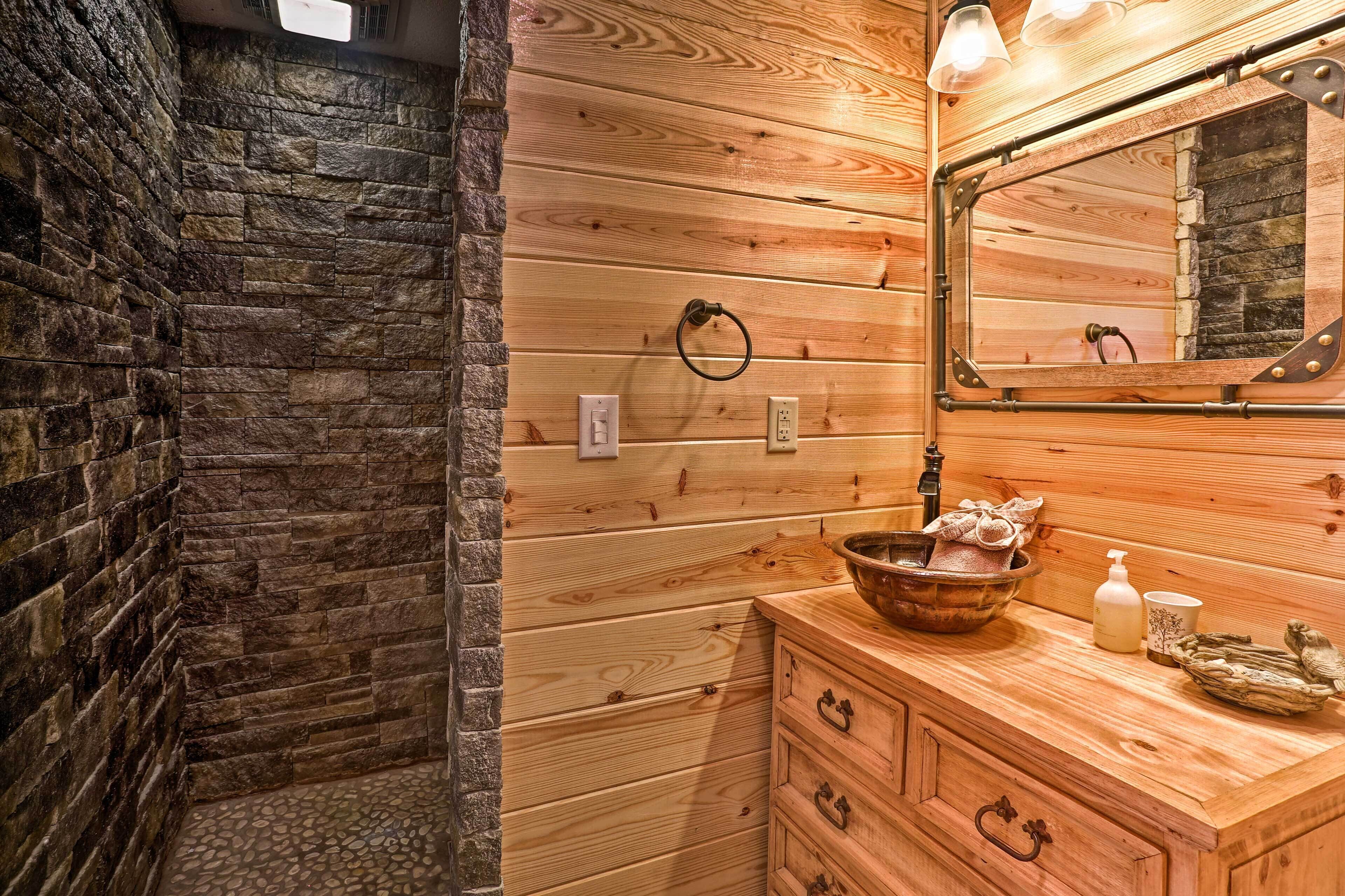 A wooden cabin bathroom with a stone wall, wooden vanity, and a mirror with a rustic frame.
