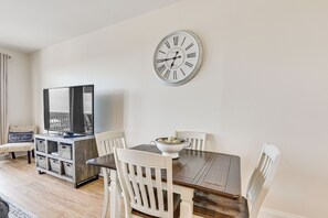 Een eetruimte met een houten tafel, witte stoelen, een grote wandklok, een televisie op een dressoir en een mand met fruit.