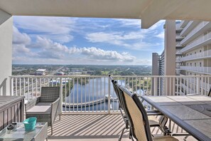 A balcony with a view of a lake and distant buildings, furnished with wicker chairs and a glass table.