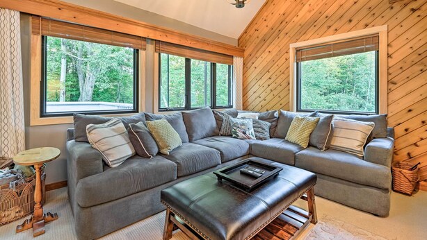 A modern living room with a sectional sofa, a coffee table, and large windows offering a view of the forest. - Family-friendly Stratton Gem Close to Slopes! (Stratton)