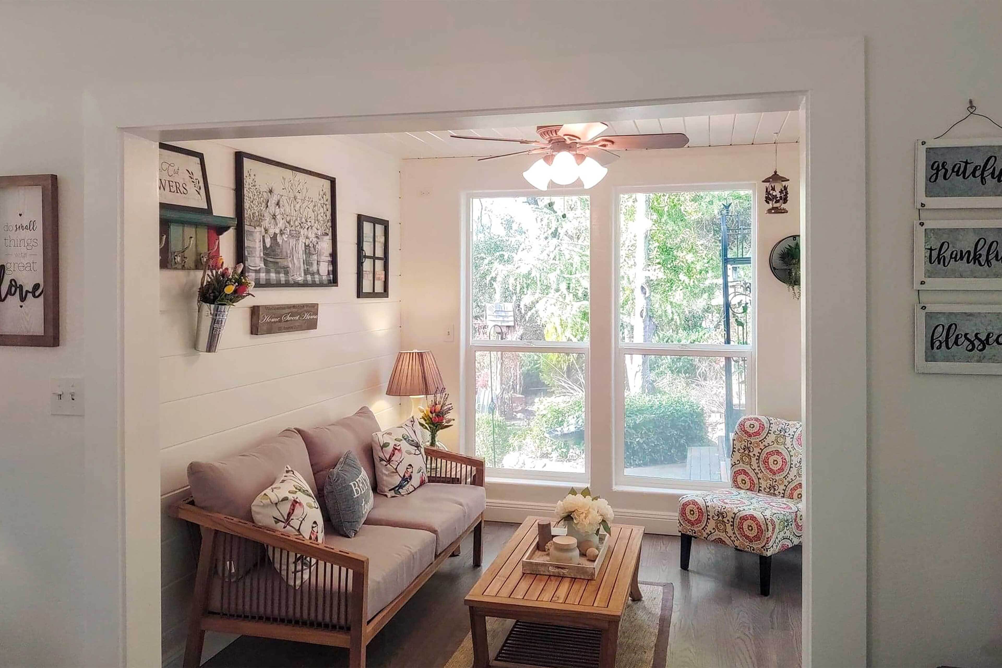 A cozy living room with a sofa, armchair, and coffee table. There are framed pictures on the wall and a ceiling fan.