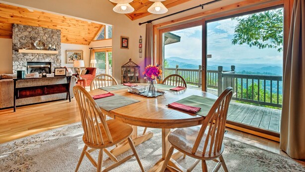 A cozy dining area with a wooden table and chairs, a stone fireplace, and a large window with a view of the mountains.