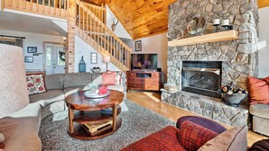 A spacious living room with a stone fireplace, wooden ceiling, and a staircase leading to an upper level.