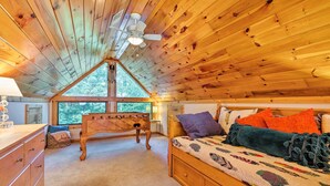 A cozy bedroom with a wooden ceiling, a bed with colorful pillows, a dresser, and a window with a view of trees.