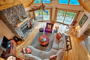 A spacious living area with a stone fireplace, a ceiling fan, and a large window overlooking a deck. - Smoky Mountain Vacation Rental Near Bryson City! (Bryson City)