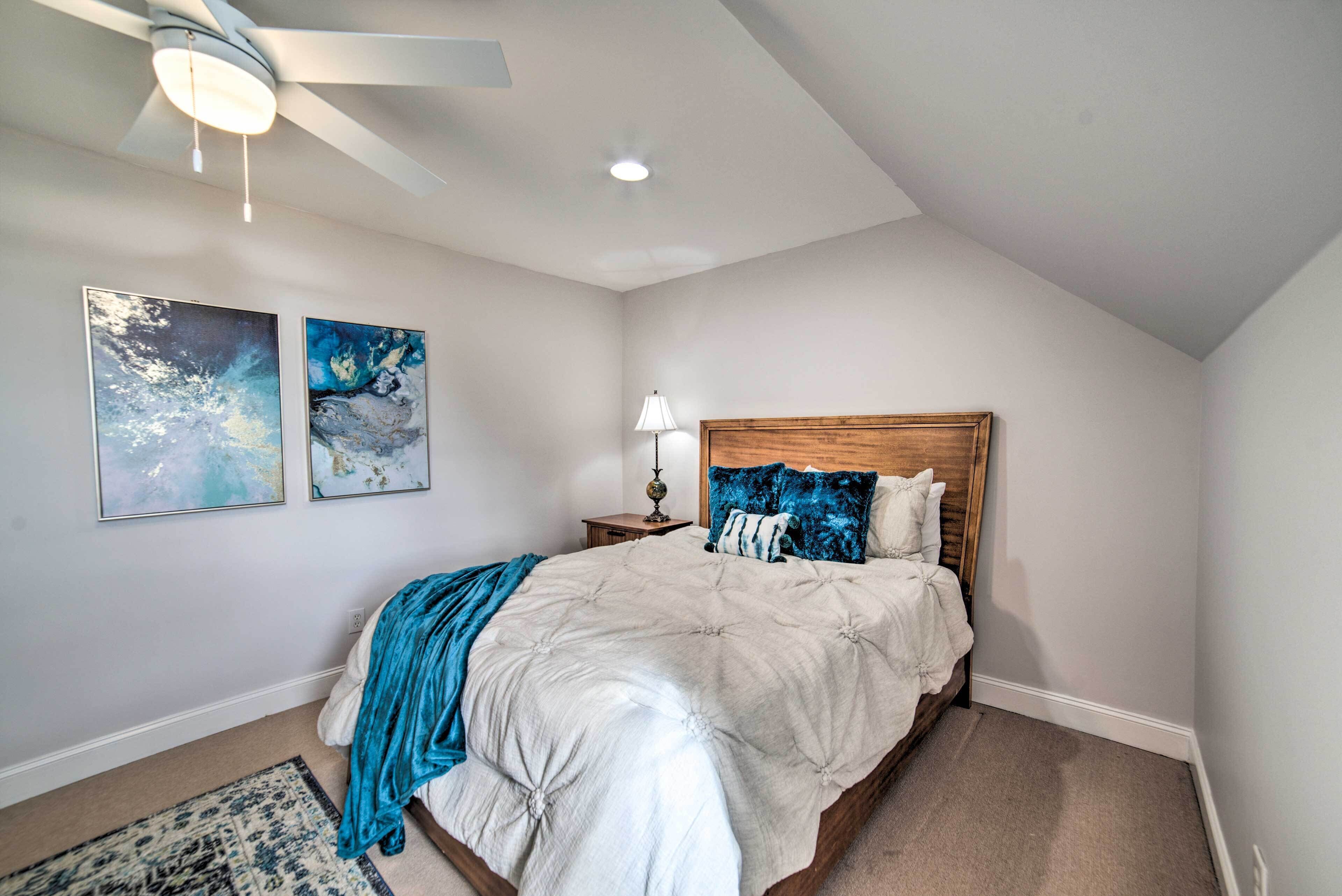 A bedroom with a bed, a ceiling fan, two wall-mounted paintings, and a nightstand with a lamp.