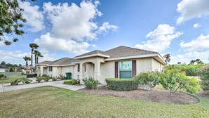 A two-story house with a brick exterior, a front yard with a lawn and shrubs, and a clear blue sky with clouds. - Laguna Vista Home w/ Resort Perks Near Beach! (Port Isabel)