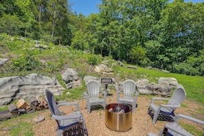 A fire pit with four Adirondack chairs arranged around it, set on a gravel surface with a backdrop of trees and rocks.