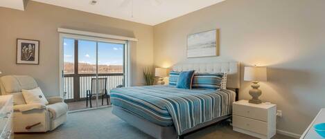 A bedroom with a large bed, a chair, a nightstand, a ceiling fan, and a view through a sliding glass door.