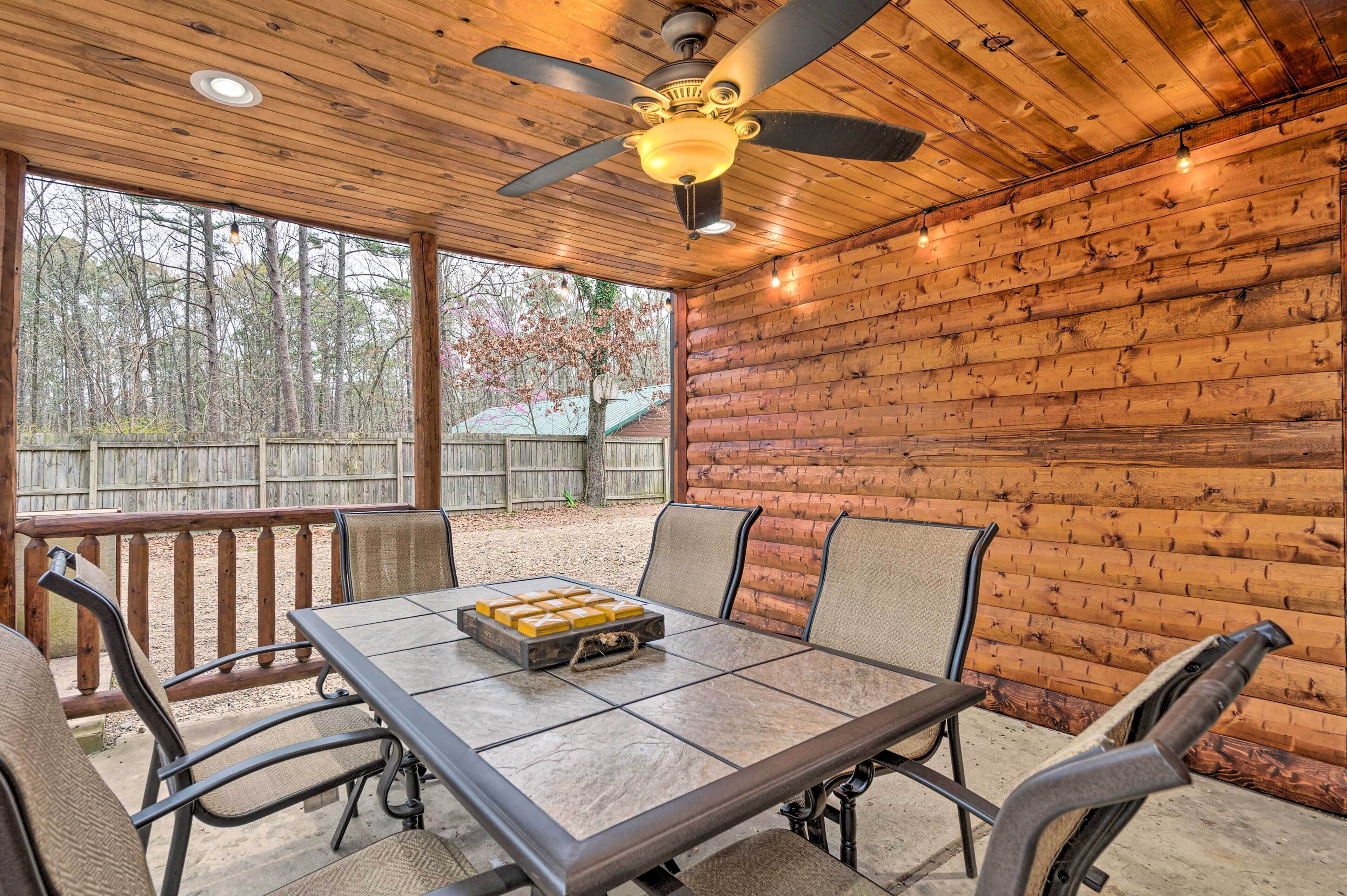 Une terrasse extérieure avec une table et des chaises, un ventilateur de plafond, et une vue sur des arbres et une clôture en bois.
